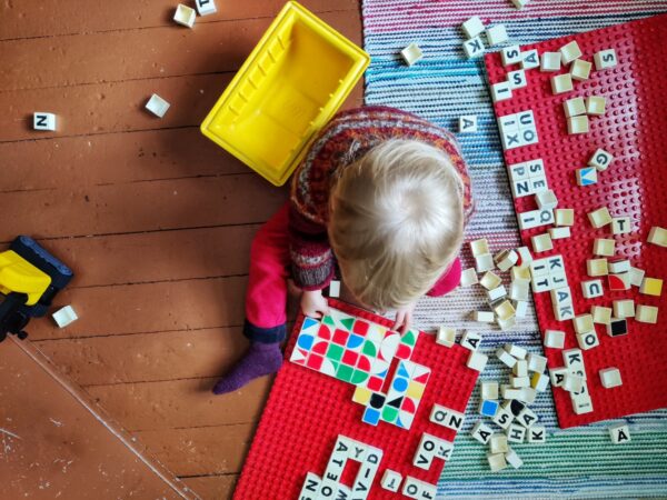 Blond-haired boy wearing home-made Norwegian sweater sits on wooden floor and plays with colorful Duplo blocks.