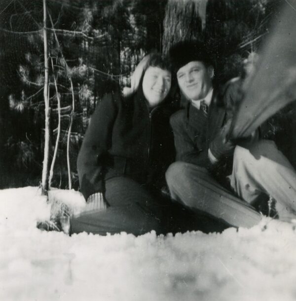 A happy couple sitting on snow, leaning against a tree, the man holding a stick towards the camera, presumably using it to capture the shot.