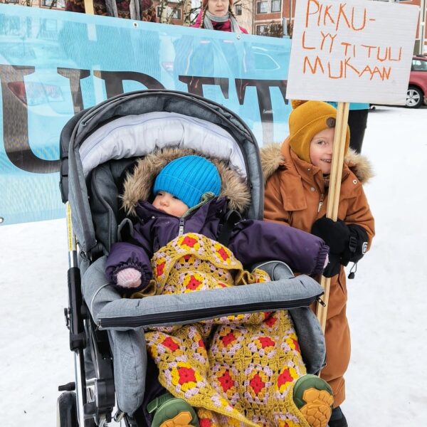 Lastenrattaissa istuvan taaperon vieressä pieni poika pitelee itse tehtyä kylttiä, jossa lukee ”Pikku-Lyyti tuli mukaan”.